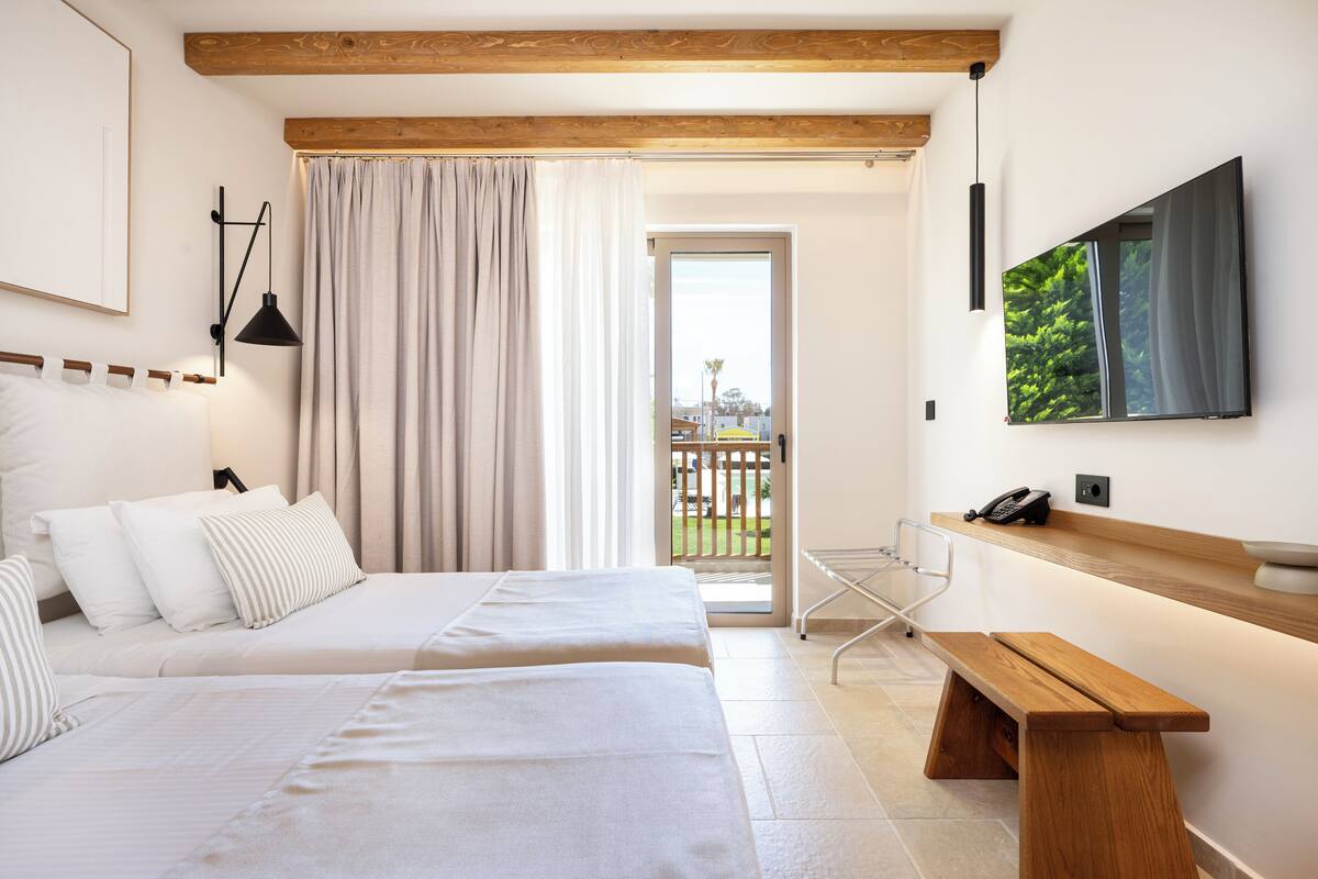 Quarto de hotel com duas camas, um banco de madeira, uma TV e uma varanda com vista.