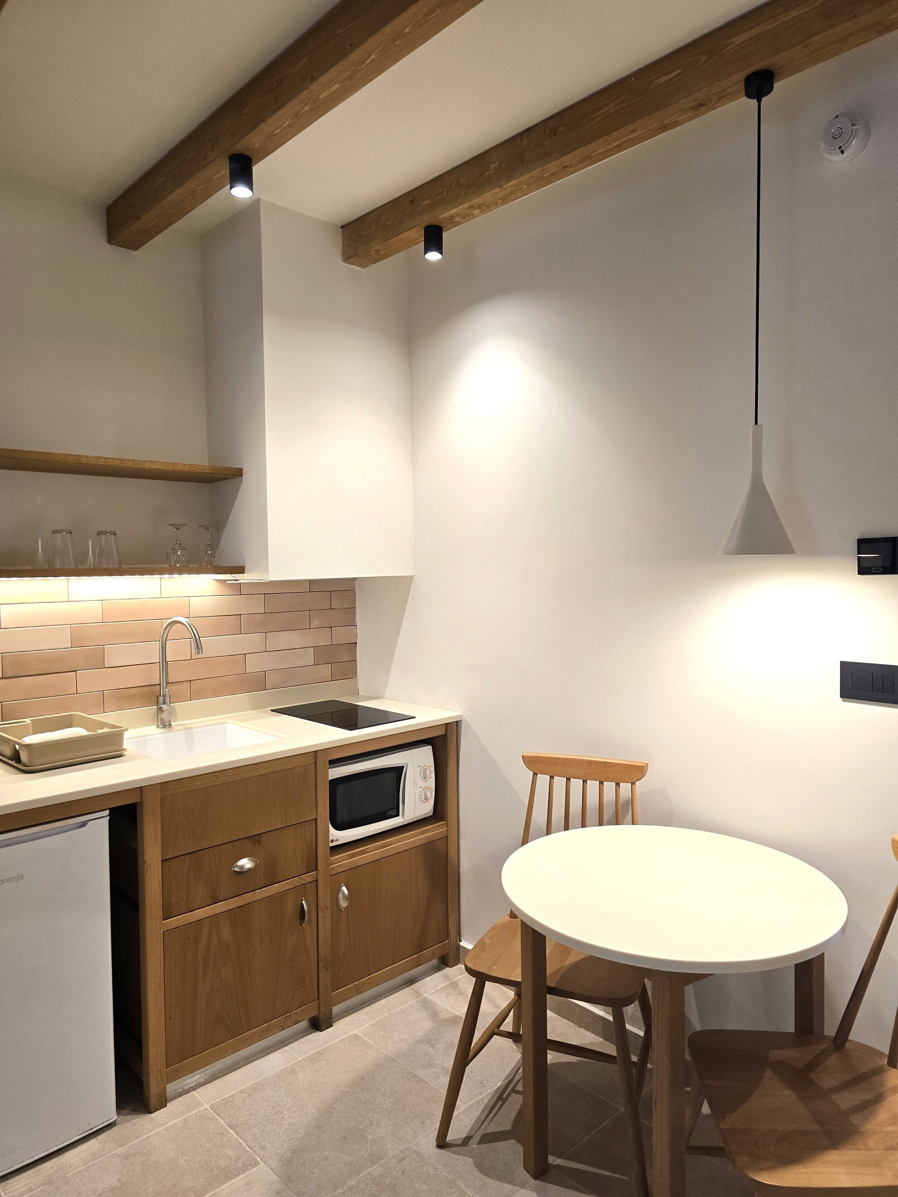 Une cuisine compacte équipée d’un évier, d’un four à micro-ondes et de meubles de rangement en bois. On y trouve une table à manger avec des chaises, et une suspension lumineuse au-dessus.