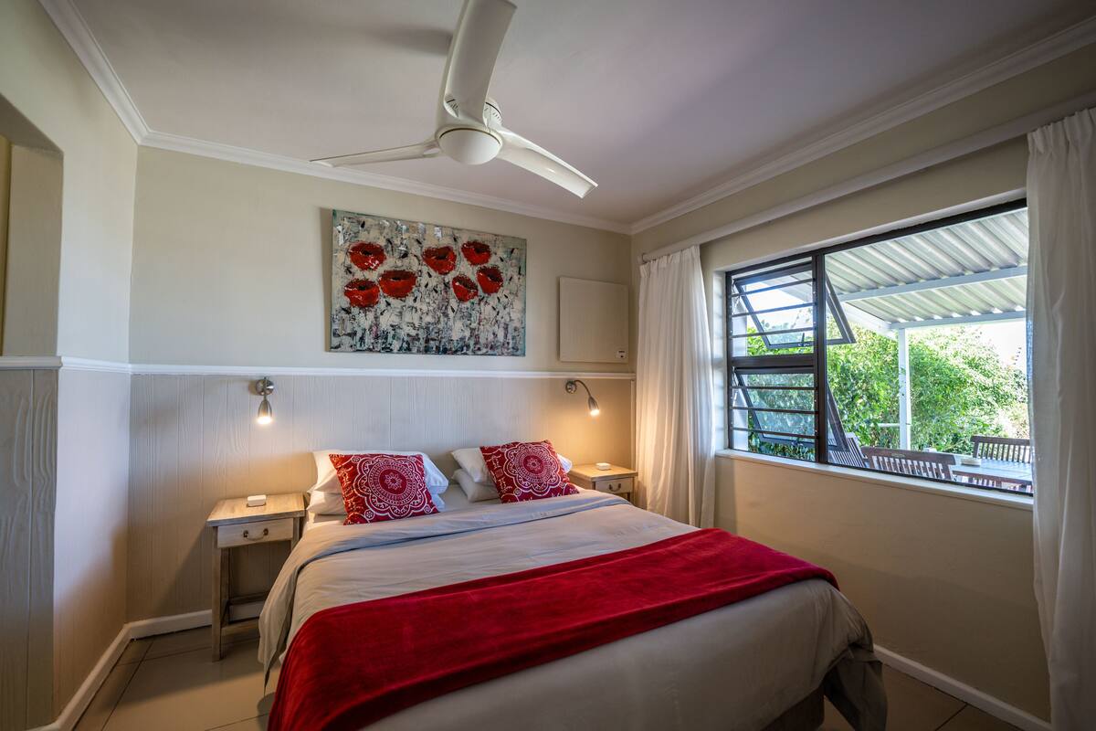 A bedroom with a bed, a ceiling fan, a nightstand, a painting, a window with curtains, and a view of greenery outside.