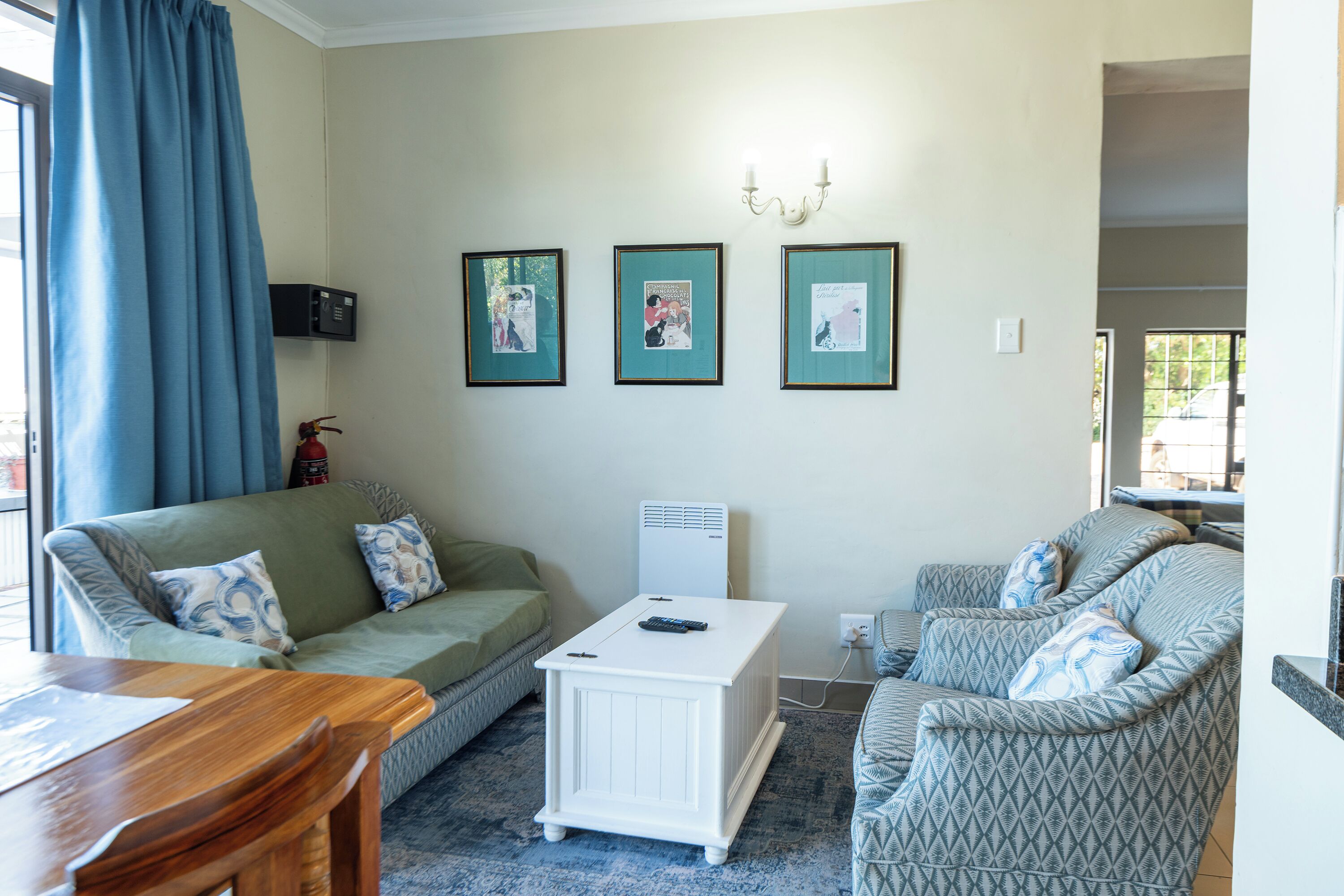 Un salon comprenant un canapé, une table basse et un fauteuil à motifs. Des tableaux encadrés ornent le mur, et un extincteur est fixé au mur.