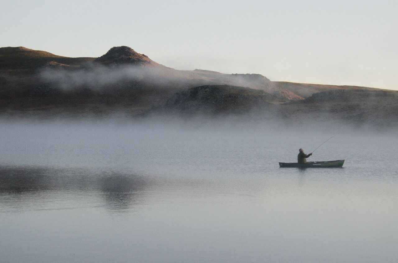Una persona pescando en un lago tranquilo con colinas cubiertas de niebla al fondo.