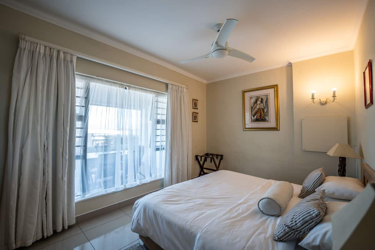A hotel room with a bed, a ceiling fan, a window with curtains, a chair, a picture on the wall, and a nightstand with a lamp.