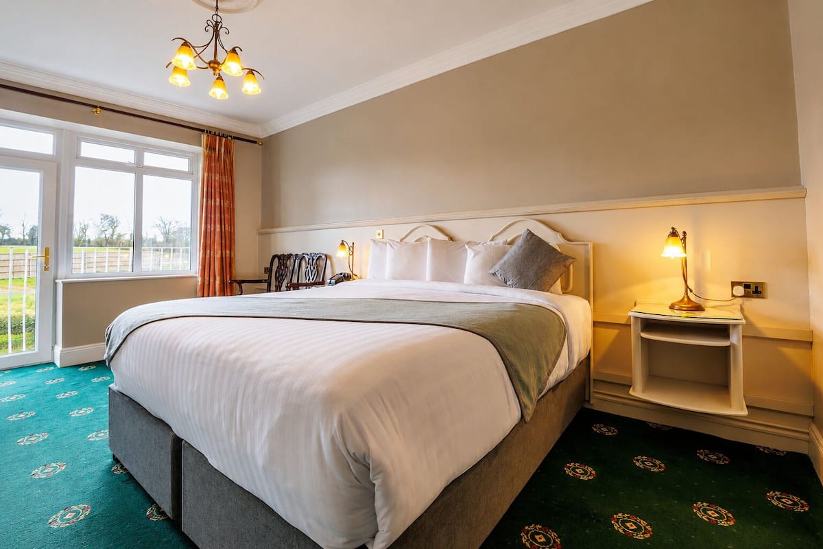 A hotel room with a large bed, a bedside table with a lamp, a window with curtains, and a view of the outdoors.