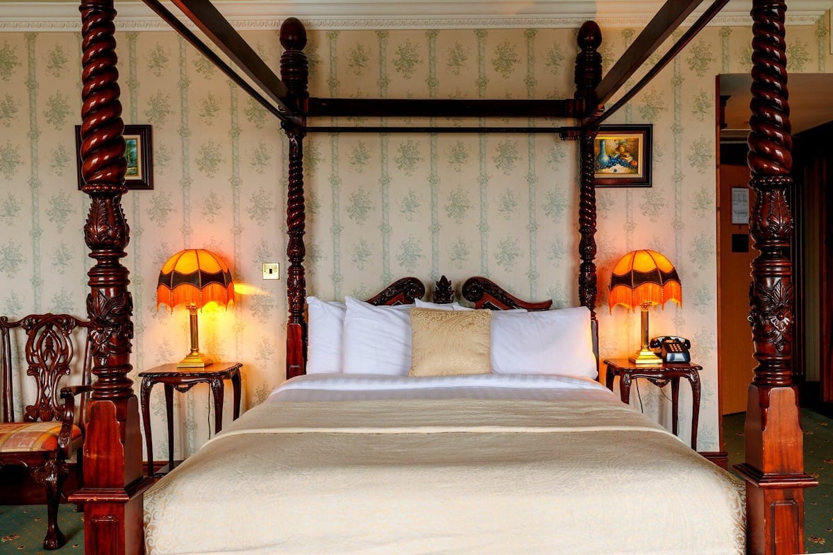 A four-poster bed with ornate wooden posts, two bedside lamps, and a floral wallpaper pattern.