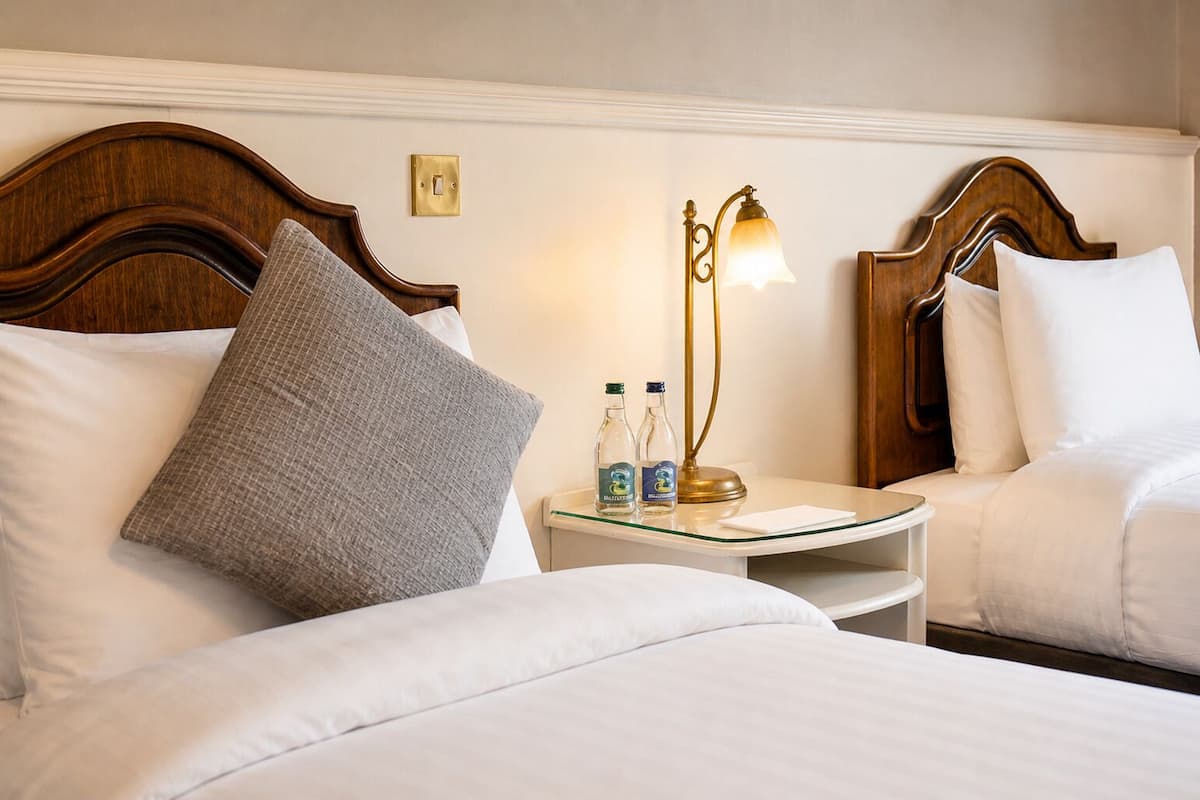 A hotel room with two beds, a nightstand with a lamp, and two bottles of water.