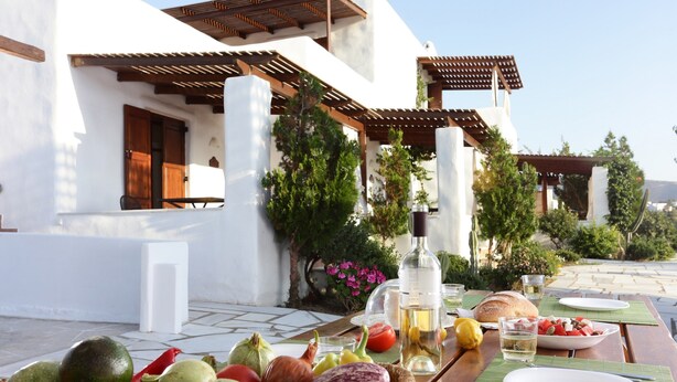 Un área de comedor al aire libre con una mesa lista para una comida, rodeada por un edificio blanco con pérgolas de madera y follaje.