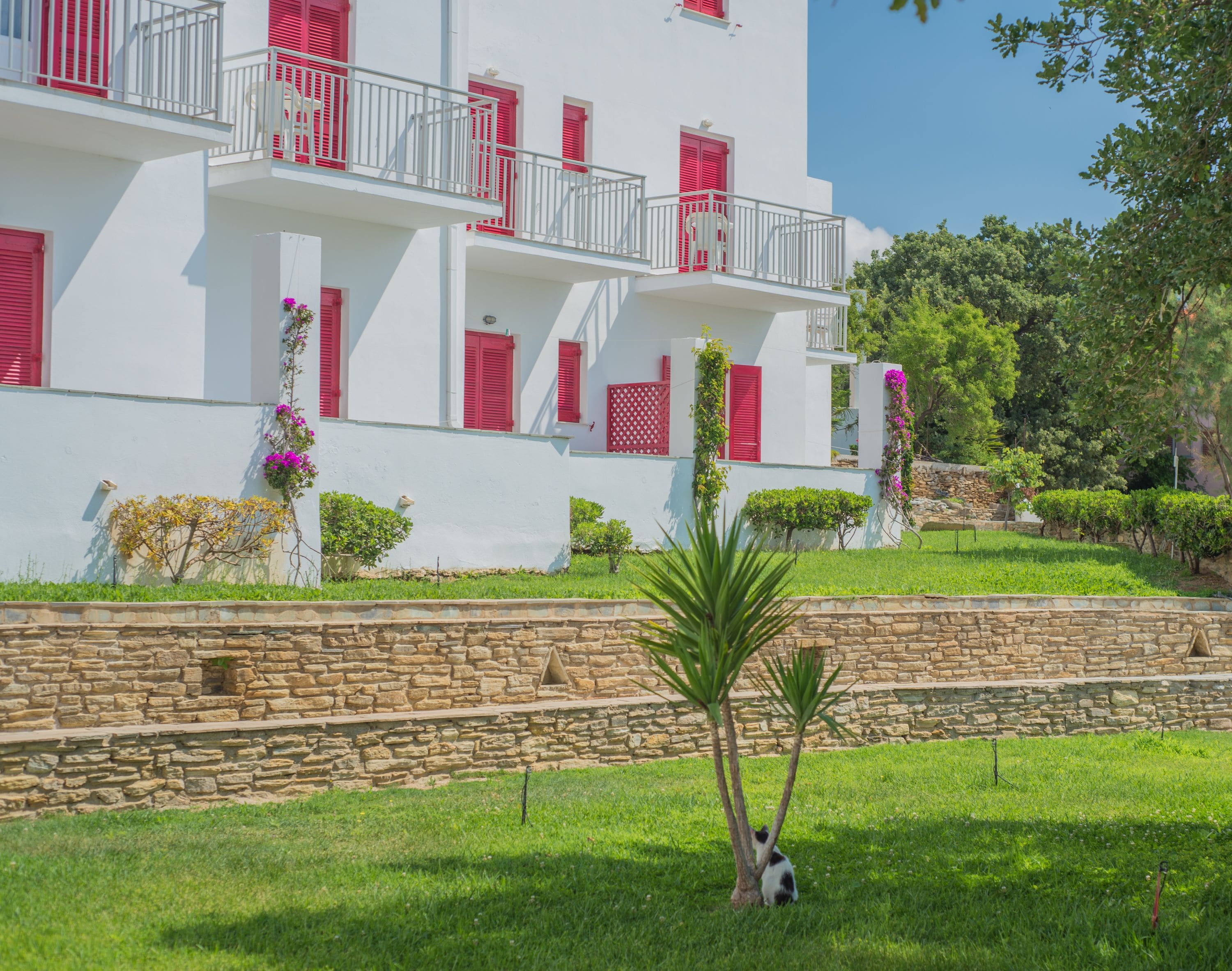 A white building with red shutters and balconies, surrounded by greenery and a stone wall.