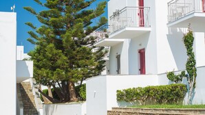 Un edificio blanco con balcones y contraventanas rojas, rodeado de vegetación y un cielo azul despejado.
