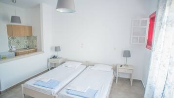 A twin room with two beds, a small bedside table with a lamp, a nightstand, a window with a red frame, and a kitchen area with a counter and cabinets.