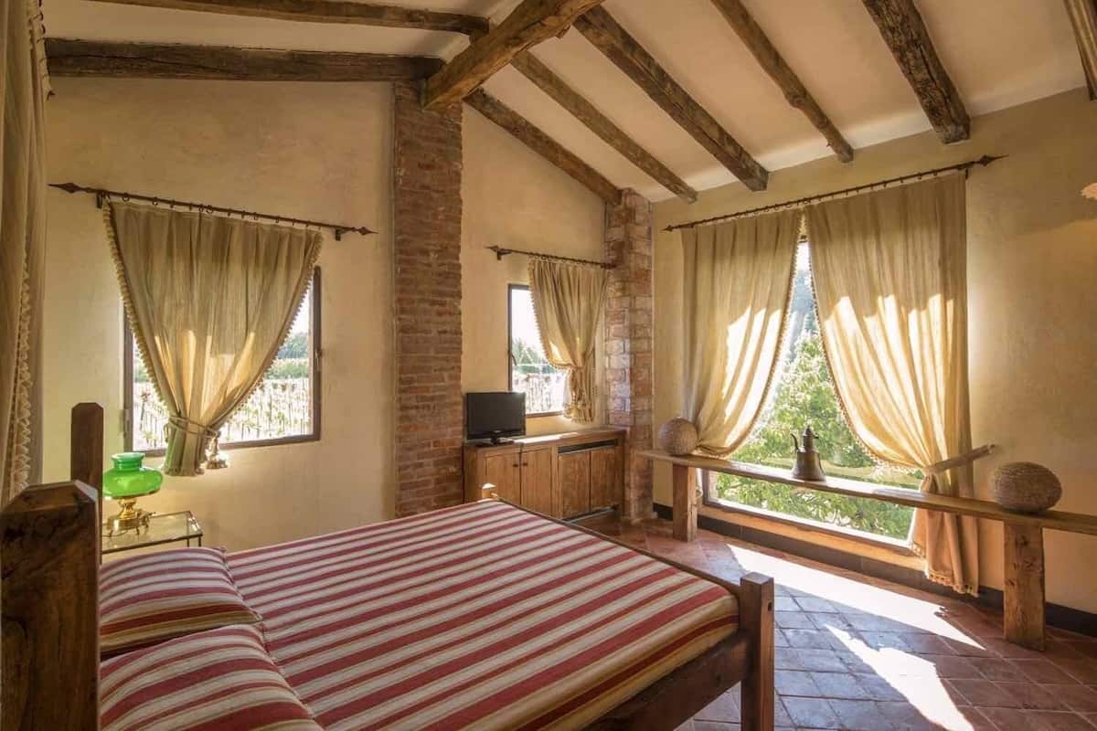 A rustic room with exposed wooden beams, a bed with striped bedding, a television on a wooden cabinet, and a view of greenery through large windows.