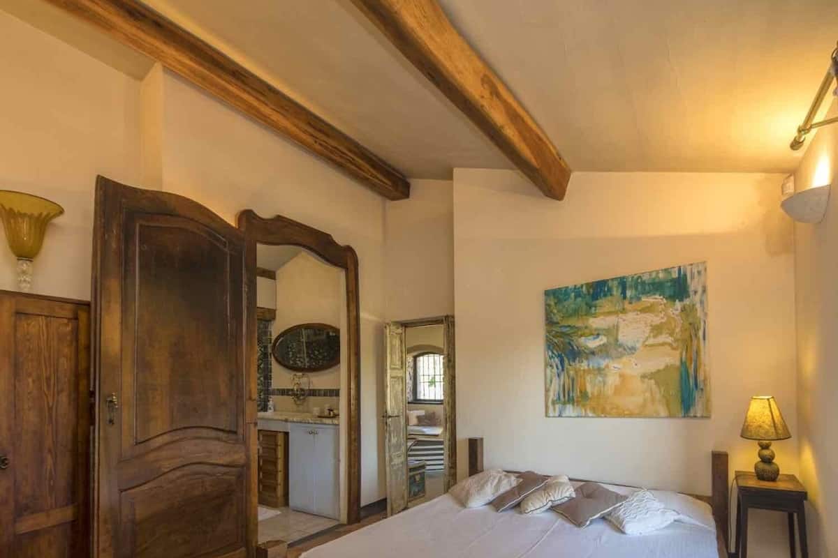 A bedroom with a wooden bed, a wooden wardrobe, a mirror, a painting on the wall, and a lamp on a side table.