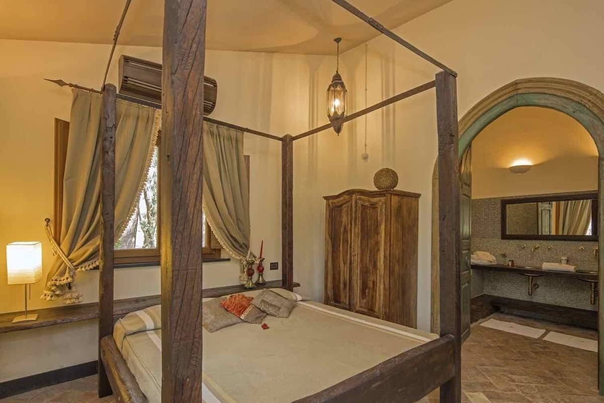 A four-poster bed with a wooden frame, a nightstand with a lamp, a mirror above the sink, and a view of the bathroom.