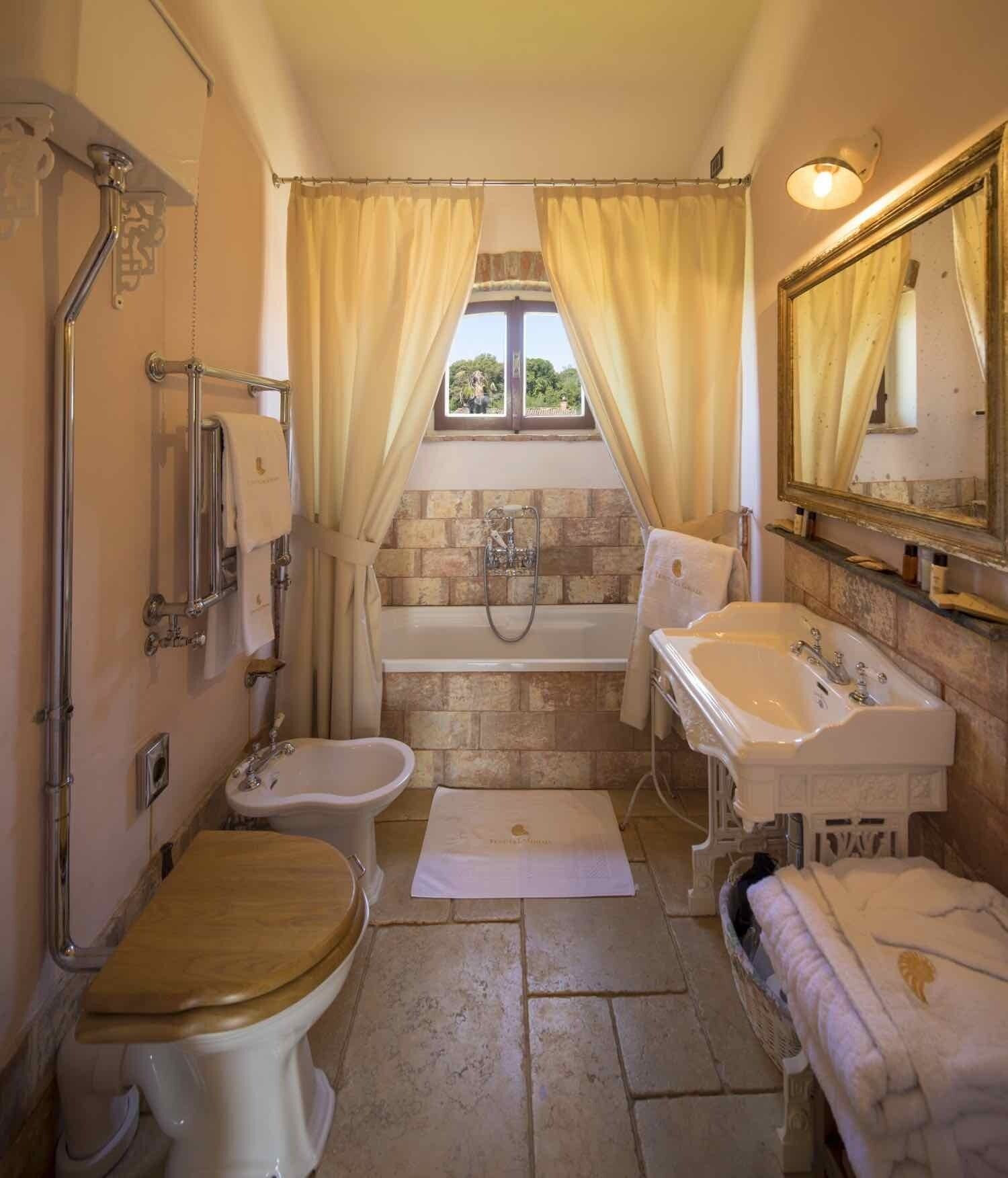 Um banheiro com banheira, vaso sanitário, bidê e pia. Há uma janela com cortinas, um espelho e um porta-toalhas.