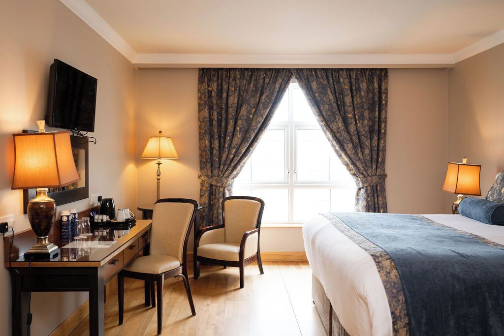 A hotel room with a bed, two chairs, a desk with a lamp, a television, and a window with curtains.