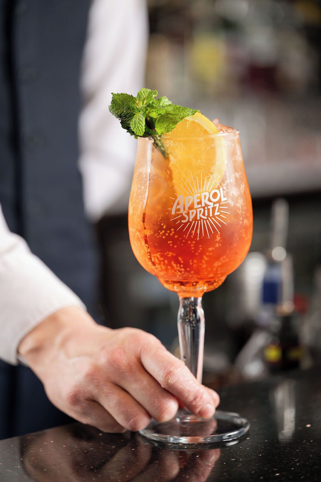 Ein Barkeeper hält ein Cocktailglas mit Zitronenscheibe und Minze, beschriftet als 'Aperol Spritz'.