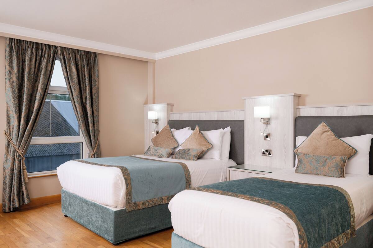 A hotel room with two beds, a window with curtains, bedside lamps, and a view of the outside.