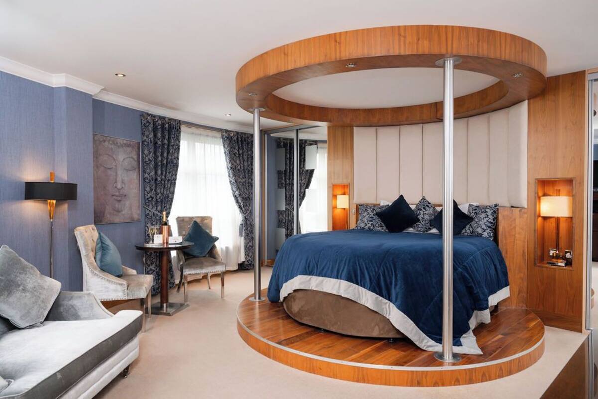 A circular bed with a wooden platform, a sofa, a small table, and a chair in a modern room with blue walls and a Buddha painting.