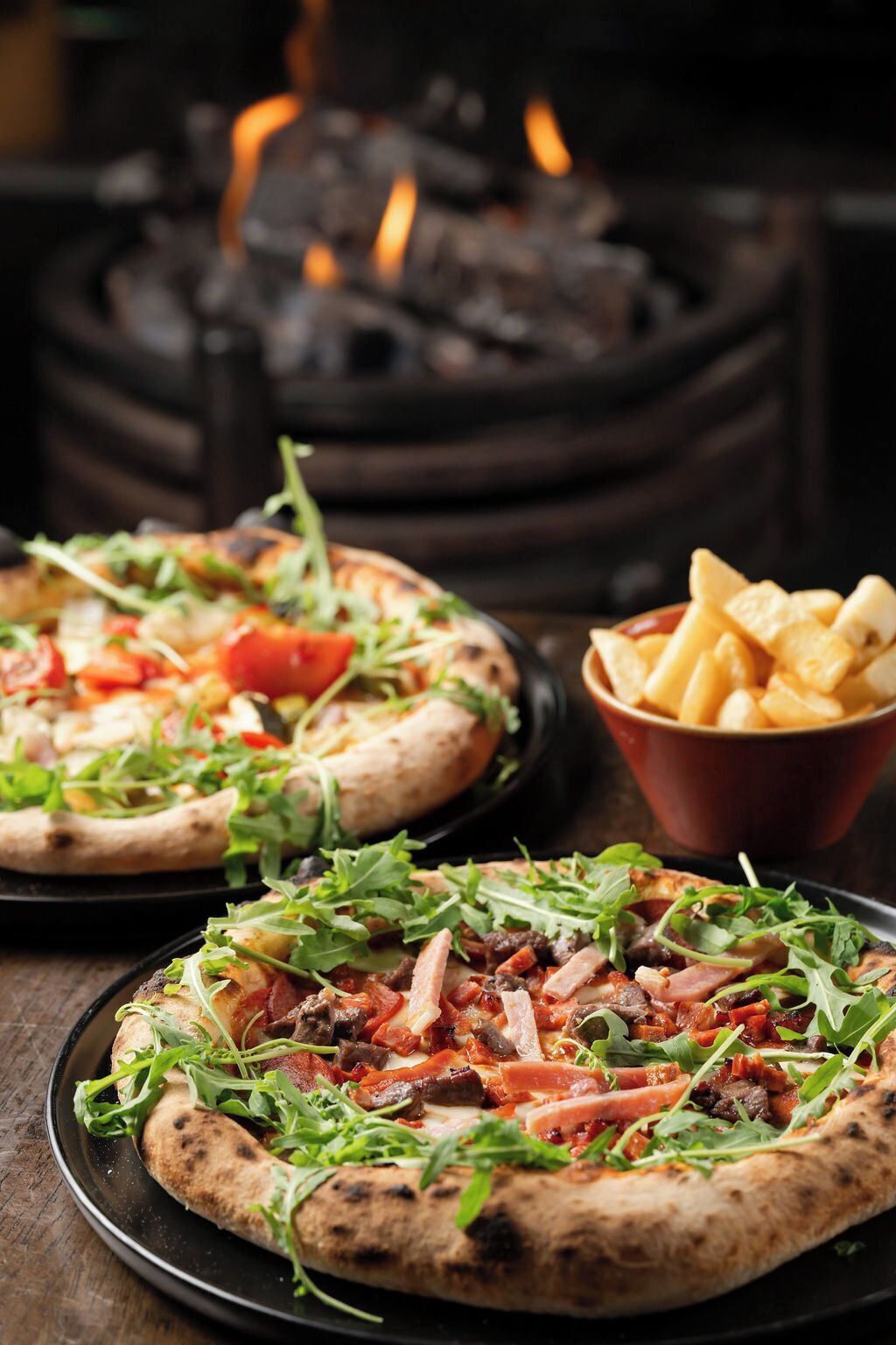 Two pizzas with various toppings on a wooden table, a bowl of fries, and a fire in the background.