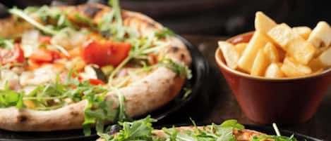 Two pizzas with various toppings on a wooden table, a bowl of fries, and a fire in the background.