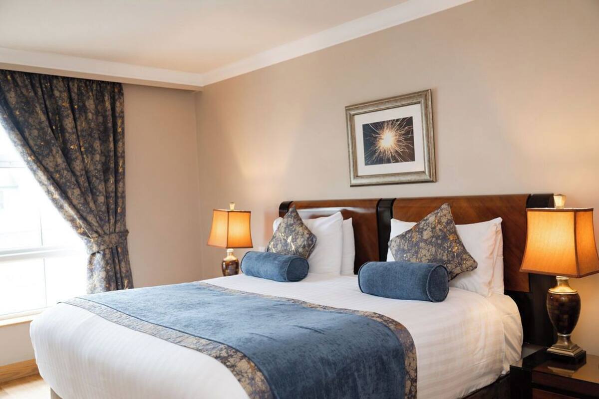A hotel room with a large bed, two bedside lamps, a framed picture on the wall, and a window with curtains.