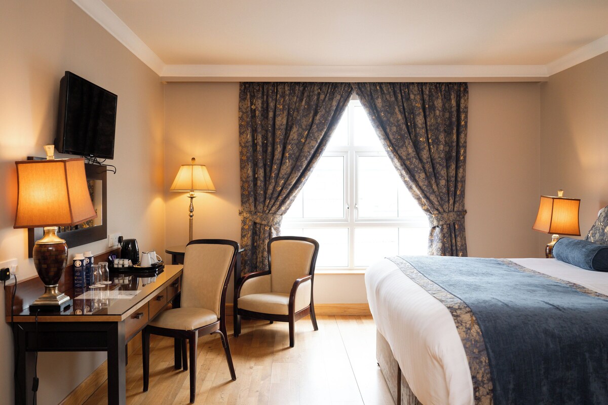 A hotel room with a bed, a desk with a lamp, a chair, a television, and a window with curtains.