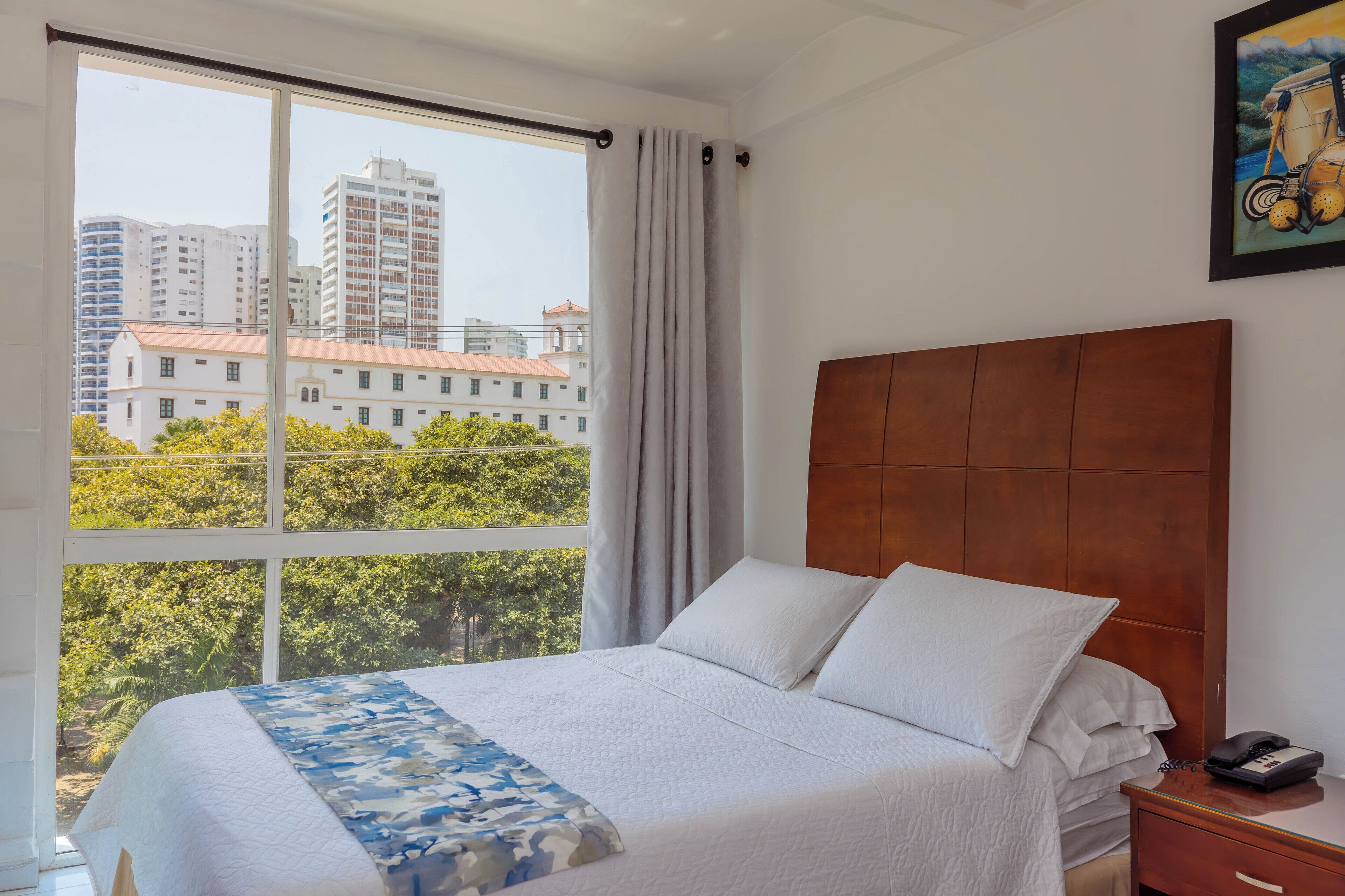 Ein Hotelzimmer mit großem Fenster, einem Bett mit weißer Bettwäsche, einem hölzernen Kopfteil und einem Nachttisch mit Telefon.