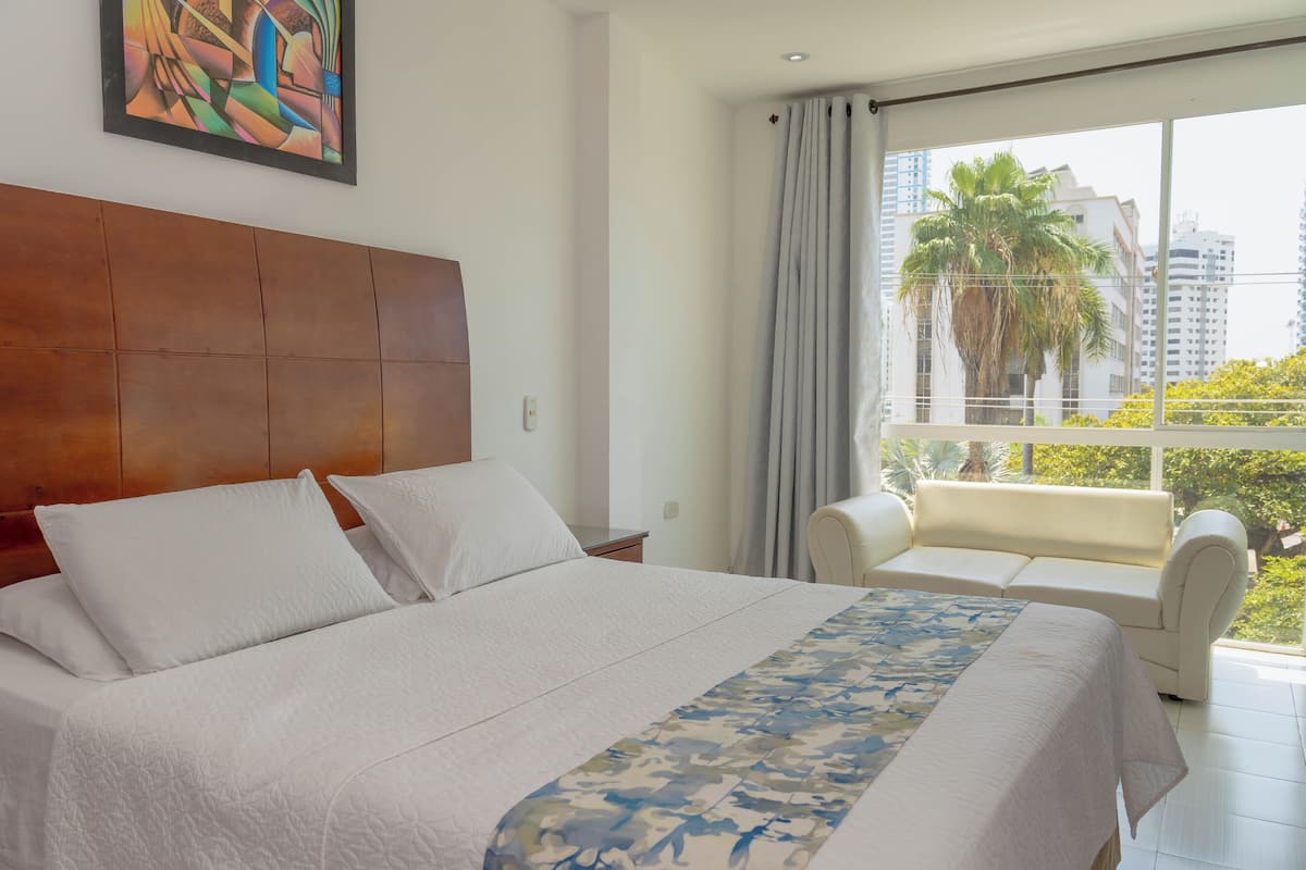 A hotel room with a large bed, a sofa, a window with a view of palm trees and buildings, and a painting on the wall.