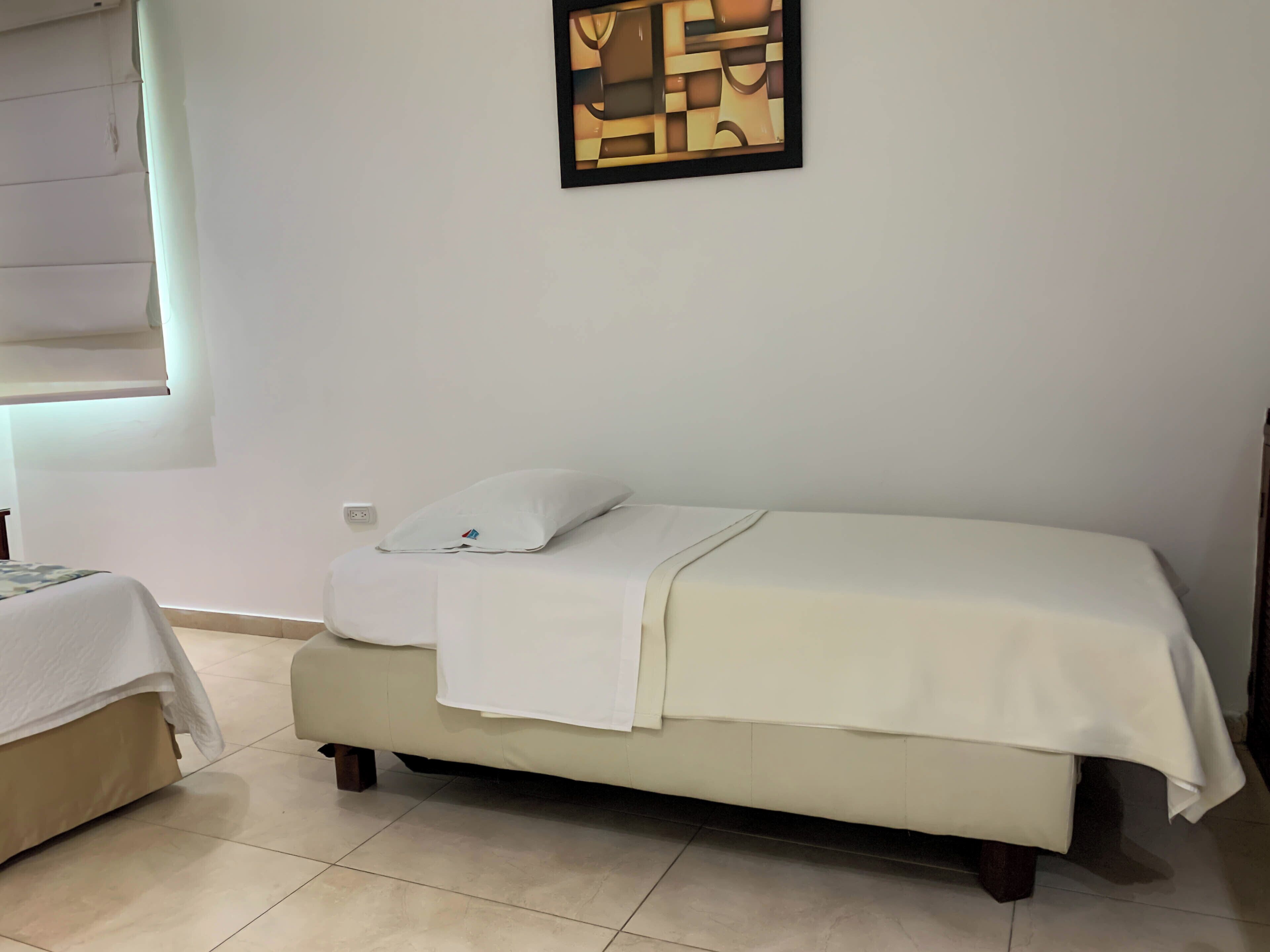 A neatly made bed with a white blanket and pillow, a small bedside table, and a wall-mounted abstract painting.