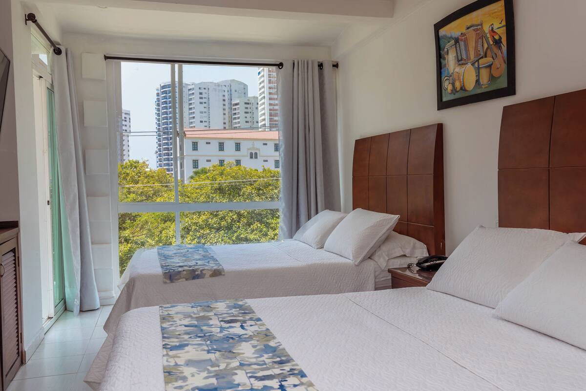 A hotel room with two beds, a large window with curtains, a wooden headboard, and a painting on the wall.