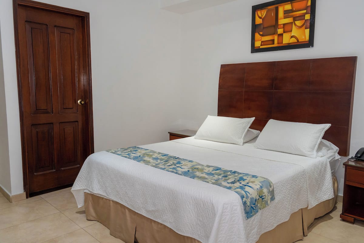A bedroom with a wooden door, a bed with white bedding, a headboard, a bedside table, and a wall-mounted artwork.