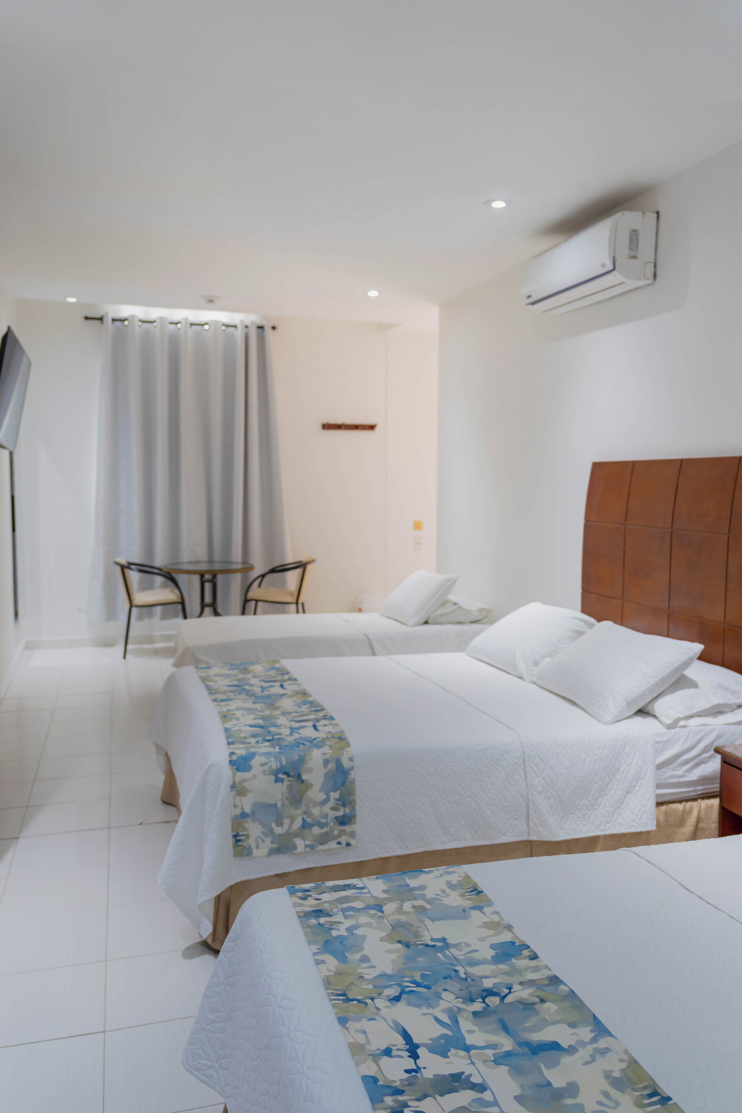 A hotel room with two beds, a small table with chairs, an air conditioning unit, and a television.