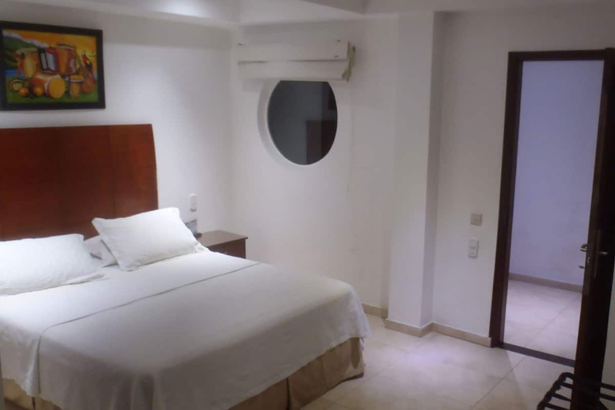 A bedroom with a bed, a round mirror, a painting, and a door leading to another room.