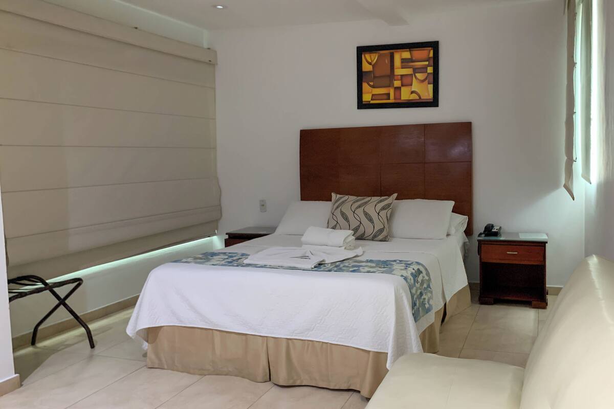 A hotel room with a bed, bedside tables, a nightstand, a chair, a window, and a wall art piece.