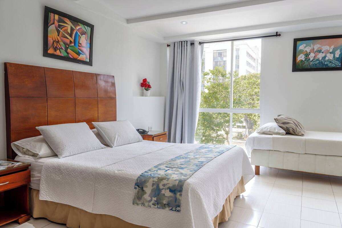 A bedroom with a large bed, a bedside table, a nightstand, a sofa, and a window with curtains.