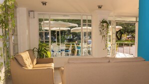 A room with a beige sofa, a window with white curtains, and a view of outdoor umbrellas and chairs. - Hotel Coral Reef Cartagena (Cartagena)