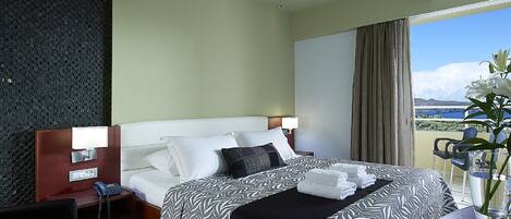 A hotel room with a bed, bedside tables, a desk, a chair, a phone, a vase with flowers, and a view of the sea.