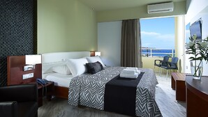 A hotel room with a bed, bedside table, chair, desk, and a view of the sea. - Eleftheria Hotel (Chania)