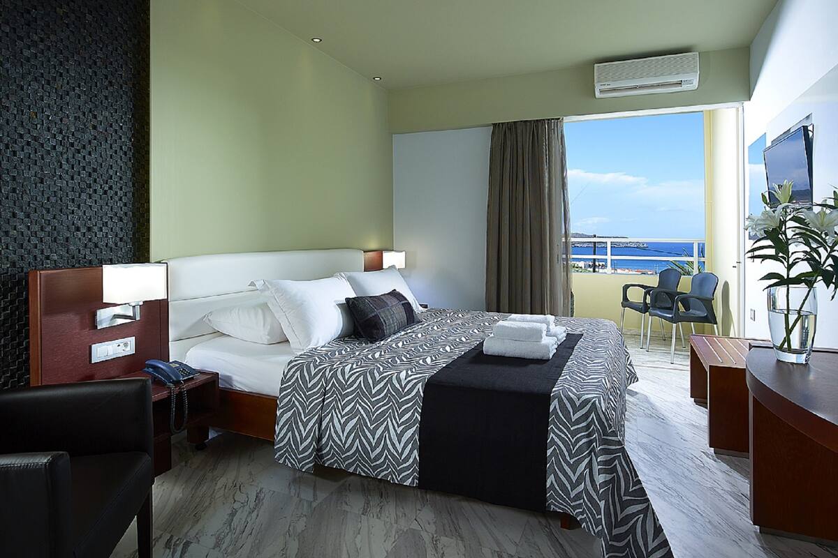 A hotel room with a bed, bedside table, chair, desk, and a view of the sea.