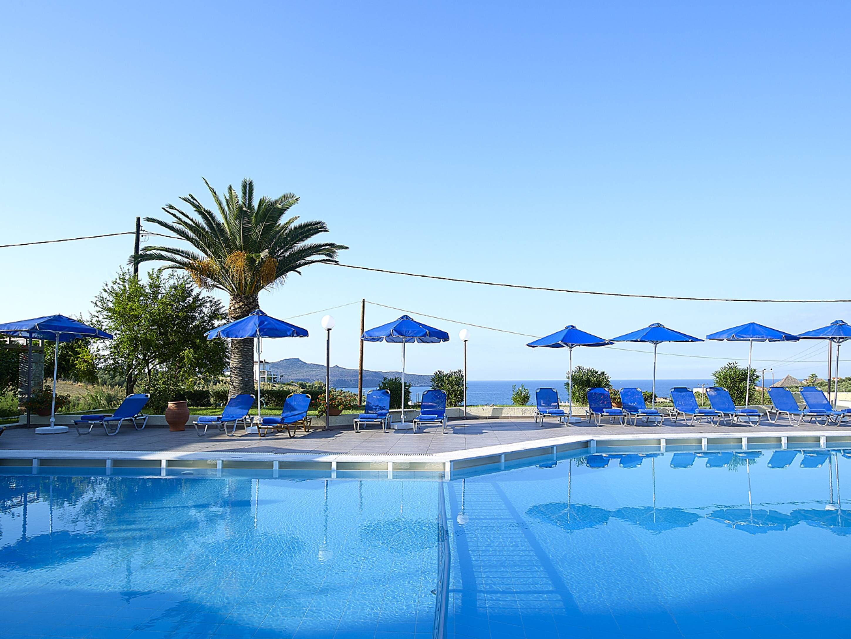 A clear blue swimming pool with lounge chairs and umbrellas, surrounded by greenery and a view of the sea.