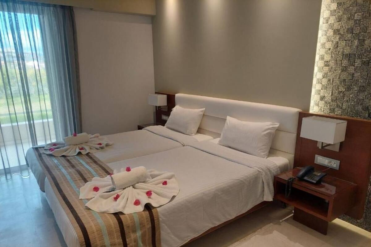A hotel room with a large bed, two bedside tables, a desk with a telephone, and a view of the outdoors through a window with curtains.