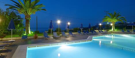 A swimming pool with lounge chairs and umbrellas, surrounded by palm trees and a view of the sea.