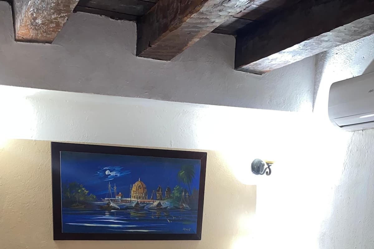 Un lit bien fait, agrémenté d’oreillers à motifs floraux et d’une couvre-lit, une tête de lit en bois, un tableau représentant un paysage côtier accroché au mur, et une petite table de chevet avec une lampe.