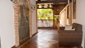 Een gang met een betegelde vloer, een muurschildering aan de linkerkant, een balkon met een balustrade, en een bank in de rechterhoek.