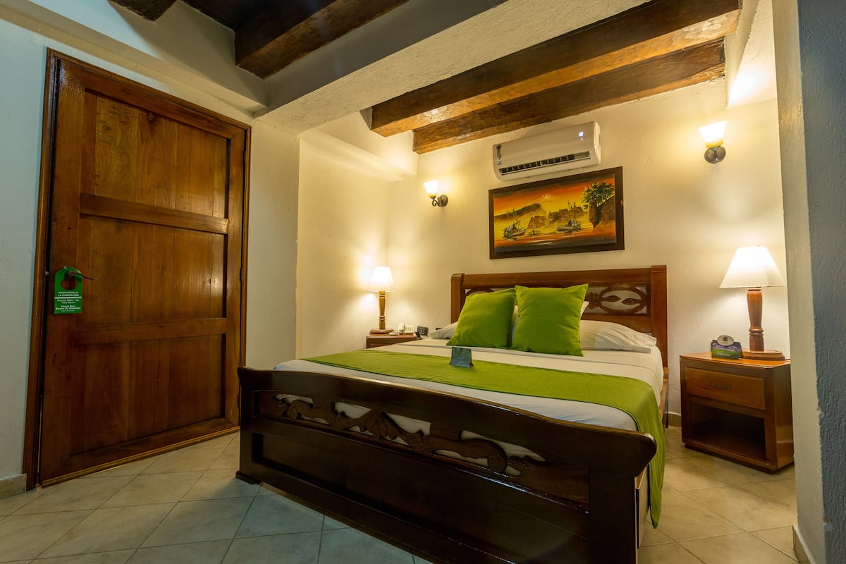 Une chambre bien rangée, avec un lit en bois, des coussins verts, deux tables de chevet avec des lampes, un tableau au mur et un appareil de climatisation.