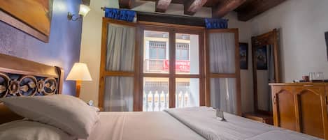 A neatly made bed with white linens, a wooden headboard with floral carvings, and a view of a balcony through a window with curtains.