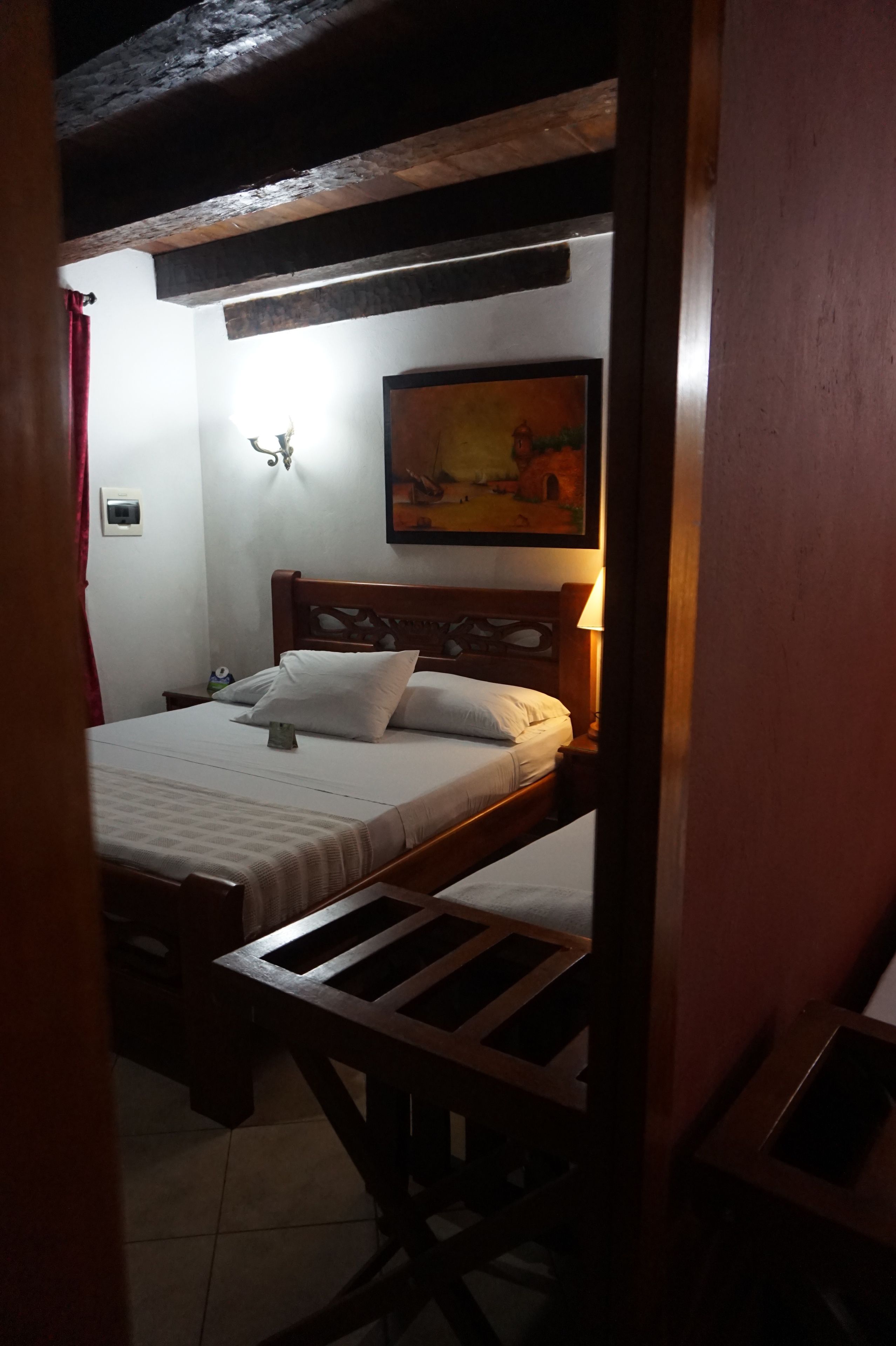 A bedroom with a wooden bed, a nightstand, a wall-mounted lamp, and a painting above the bed.