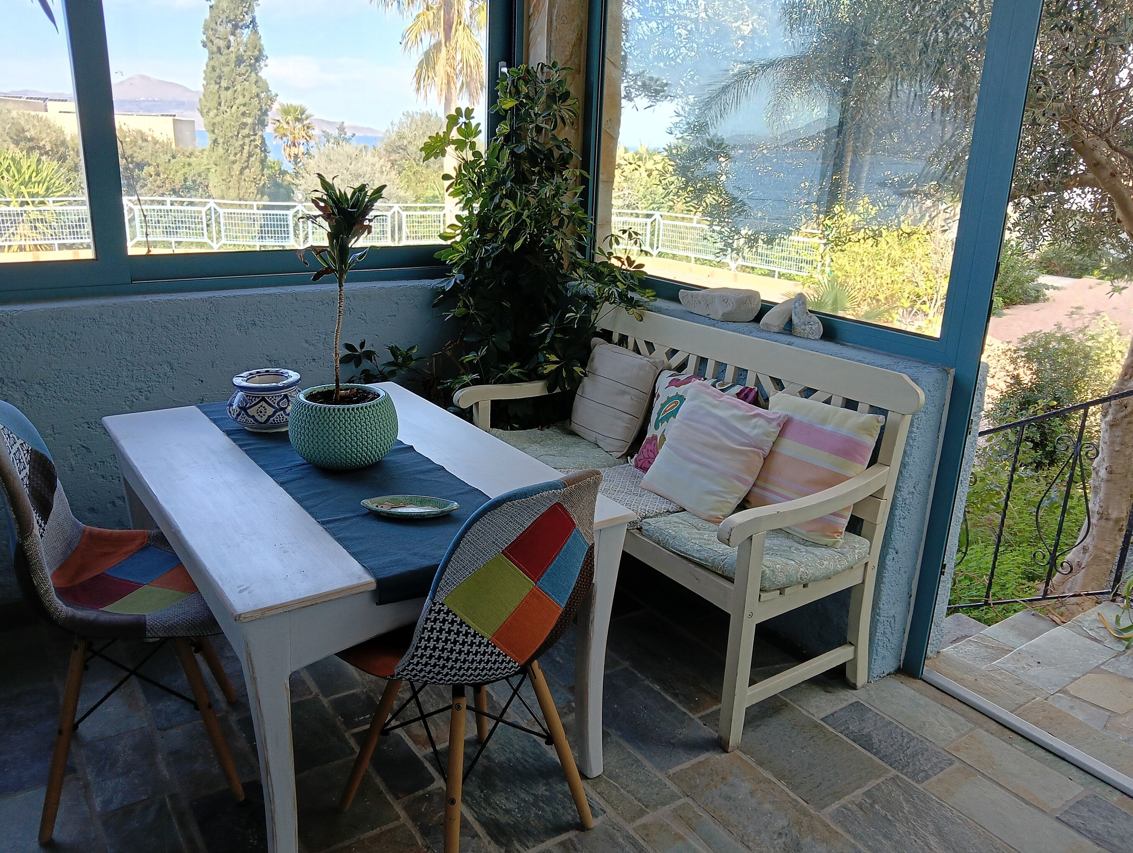 A sunlit patio with a table and chairs, a potted plant, and a bench with cushions.