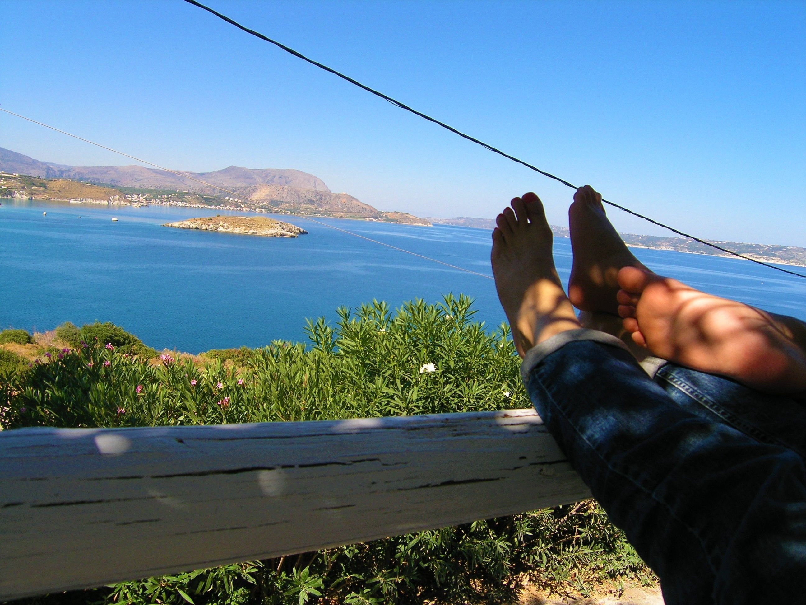 A person's feet resting on a wooden railing overlooking a coastal landscape with a clear sky.