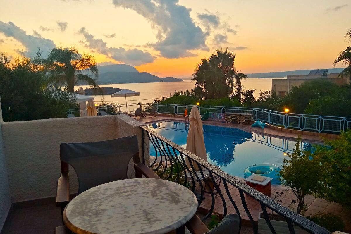 A poolside area with a view of a sunset over a body of water, surrounded by greenery and palm trees.