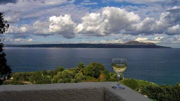 Une vue sur la côte, avec un verre de vin blanc posé sur un rebord de pierre, donnant sur une mer calme et des montagnes lointaines sous un ciel partiellement nuageux.